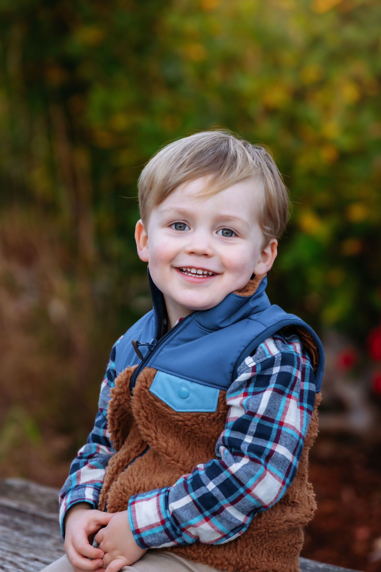 Child wearing blue vest and plaid shirt smiles outdoors against autumn foliage background.