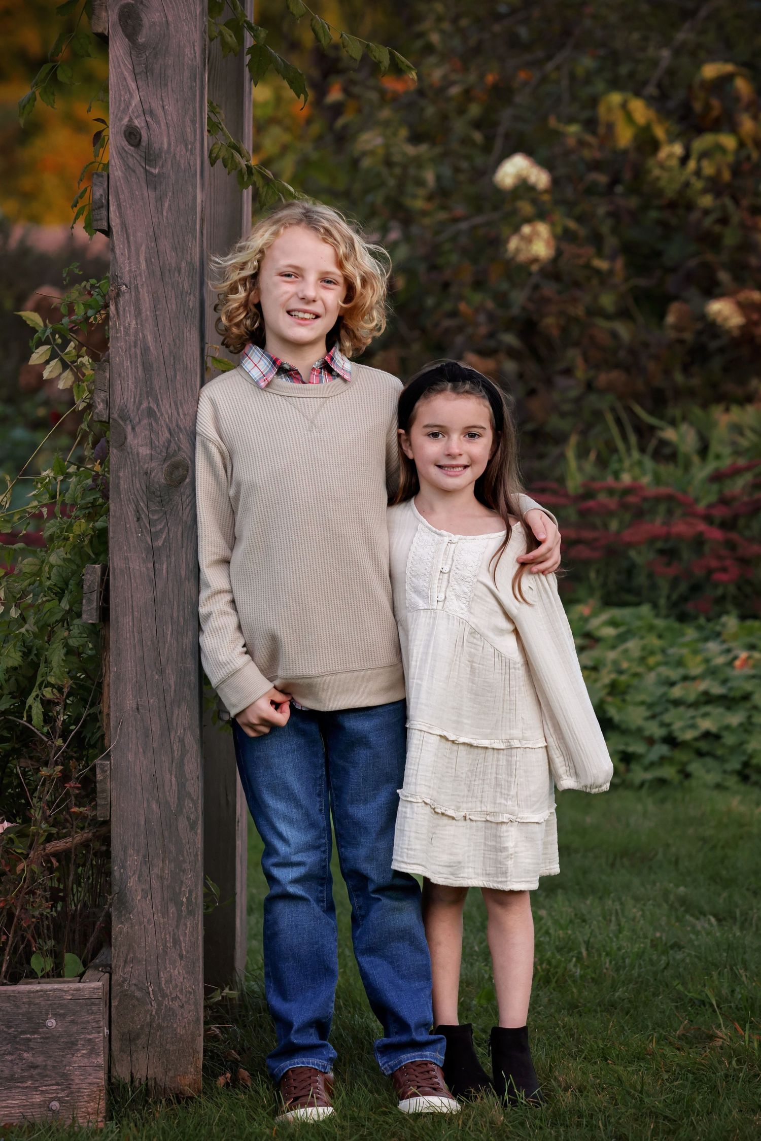 Two children stand together outdoors by a wooden post in casual fall attire.