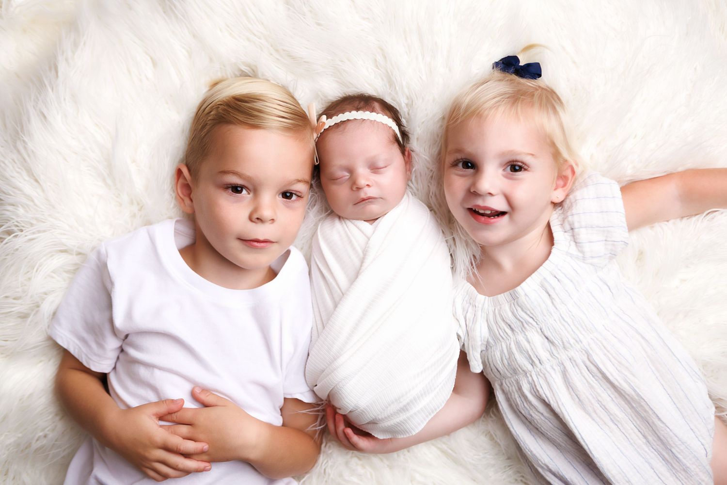 Three children in white clothing lie together on a soft white furry blanket.