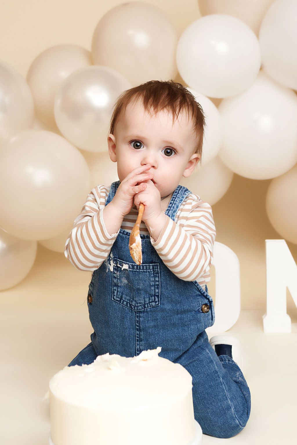 Baby in denim overalls eating cake at first birthday party with white balloons in background.