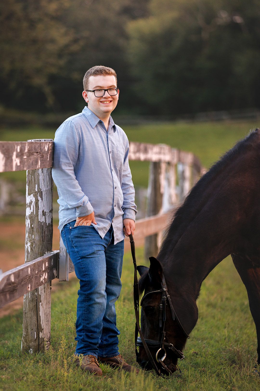 Smiling young man in glasses and blue shirt stands by wooden fence holding a dark horse grazing on green grass.