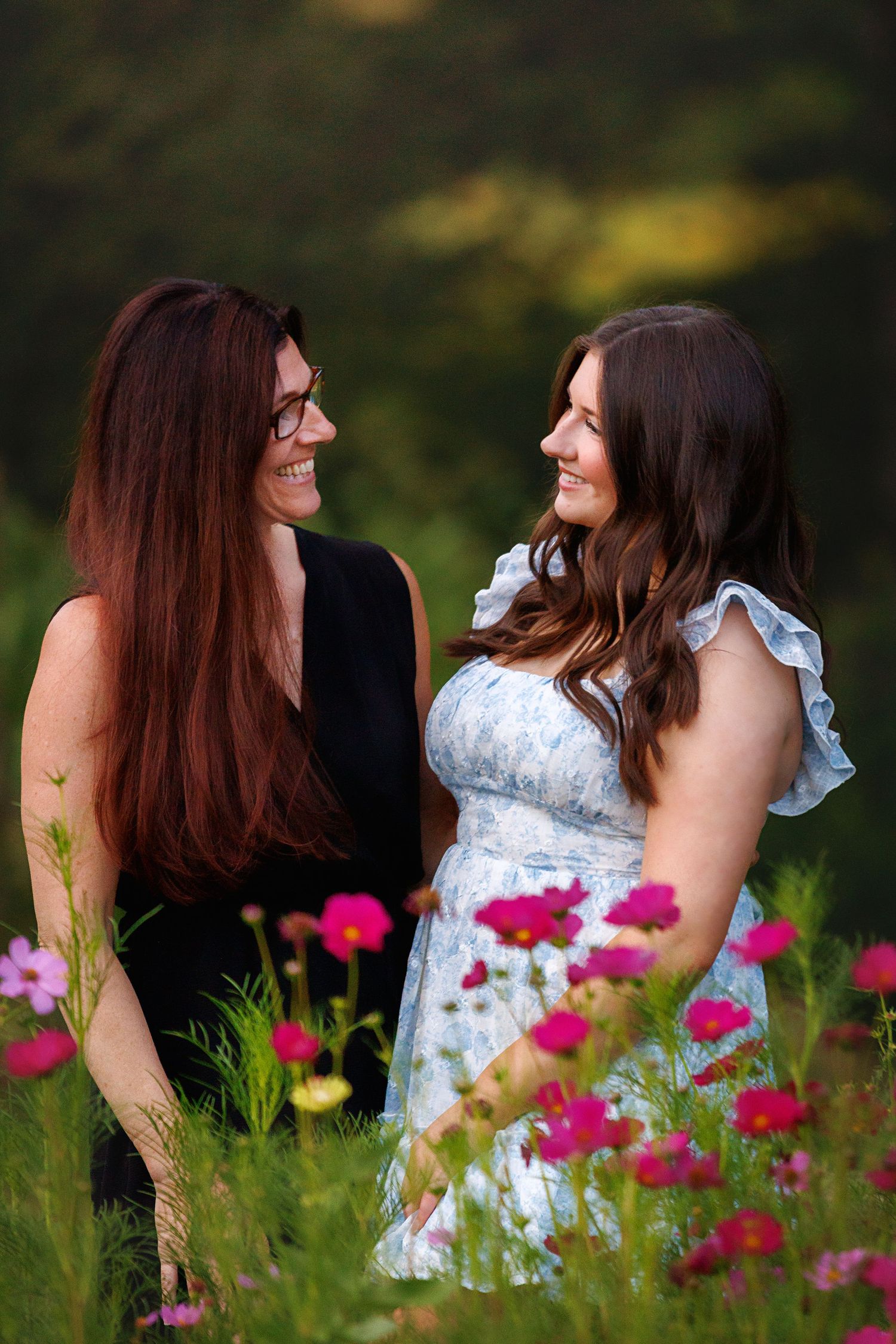 Two women smiling at each other in a field of pink cosmos flowers during golden hour.