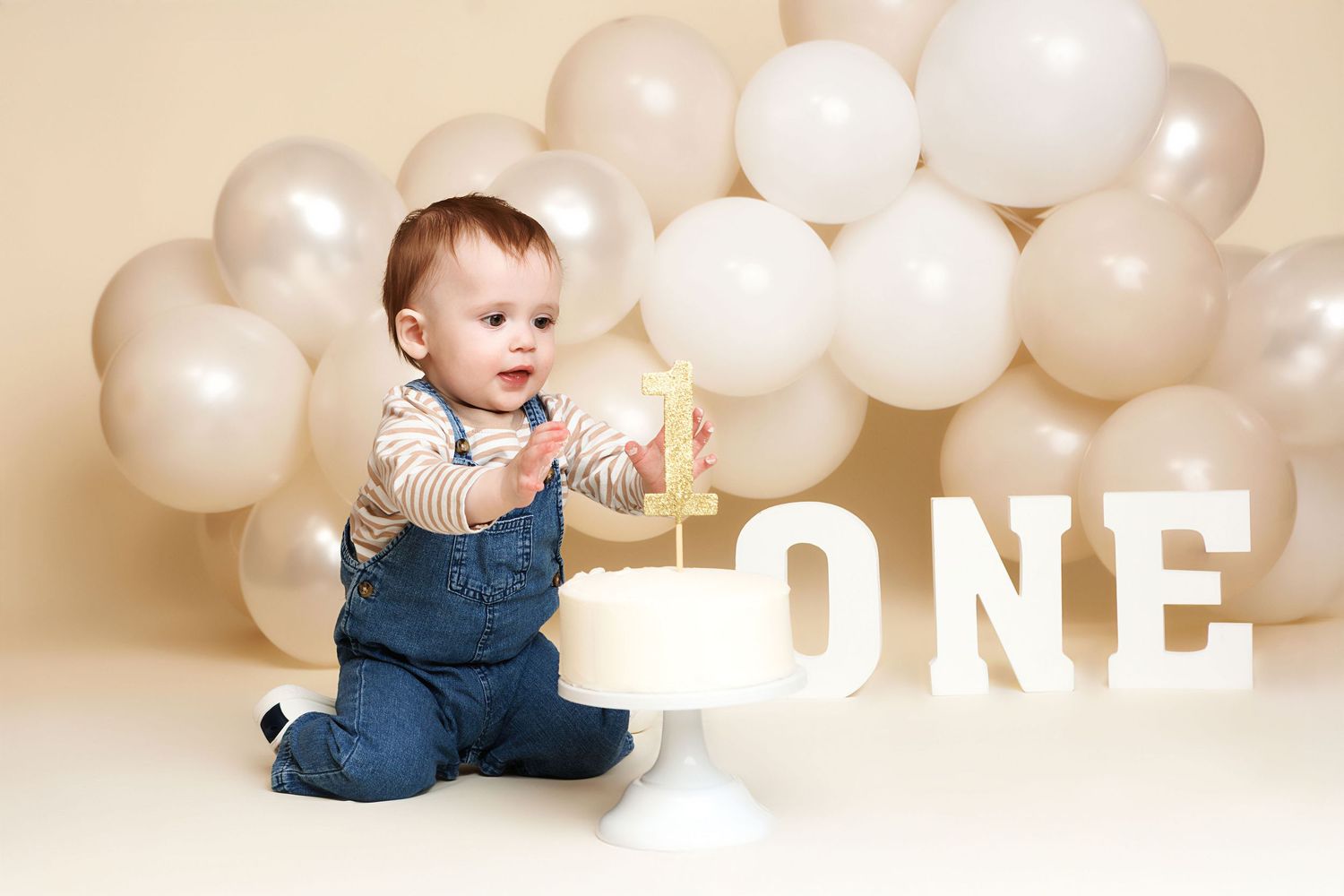 Baby boy in denim overalls sits beside a white cake at a first birthday smash cake photo session with balloons.