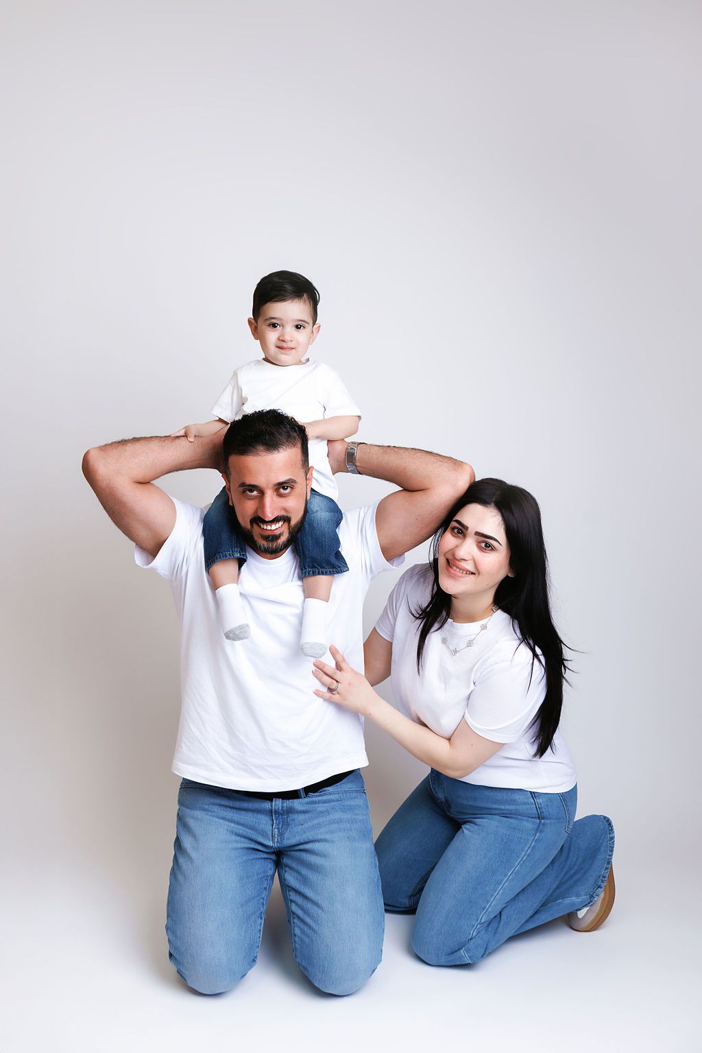 Happy family with parents in white tees and jeans posing with toddler on dad's shoulders against white background.