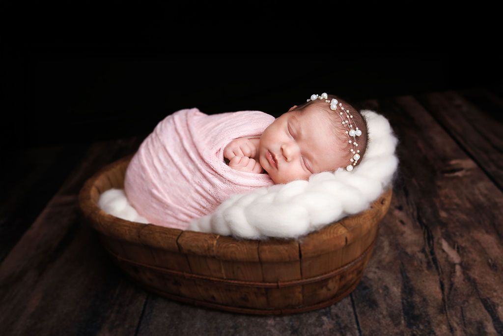 Sleeping newborn baby girl swaddled in pink wrap, resting in a wooden bowl with white fur.