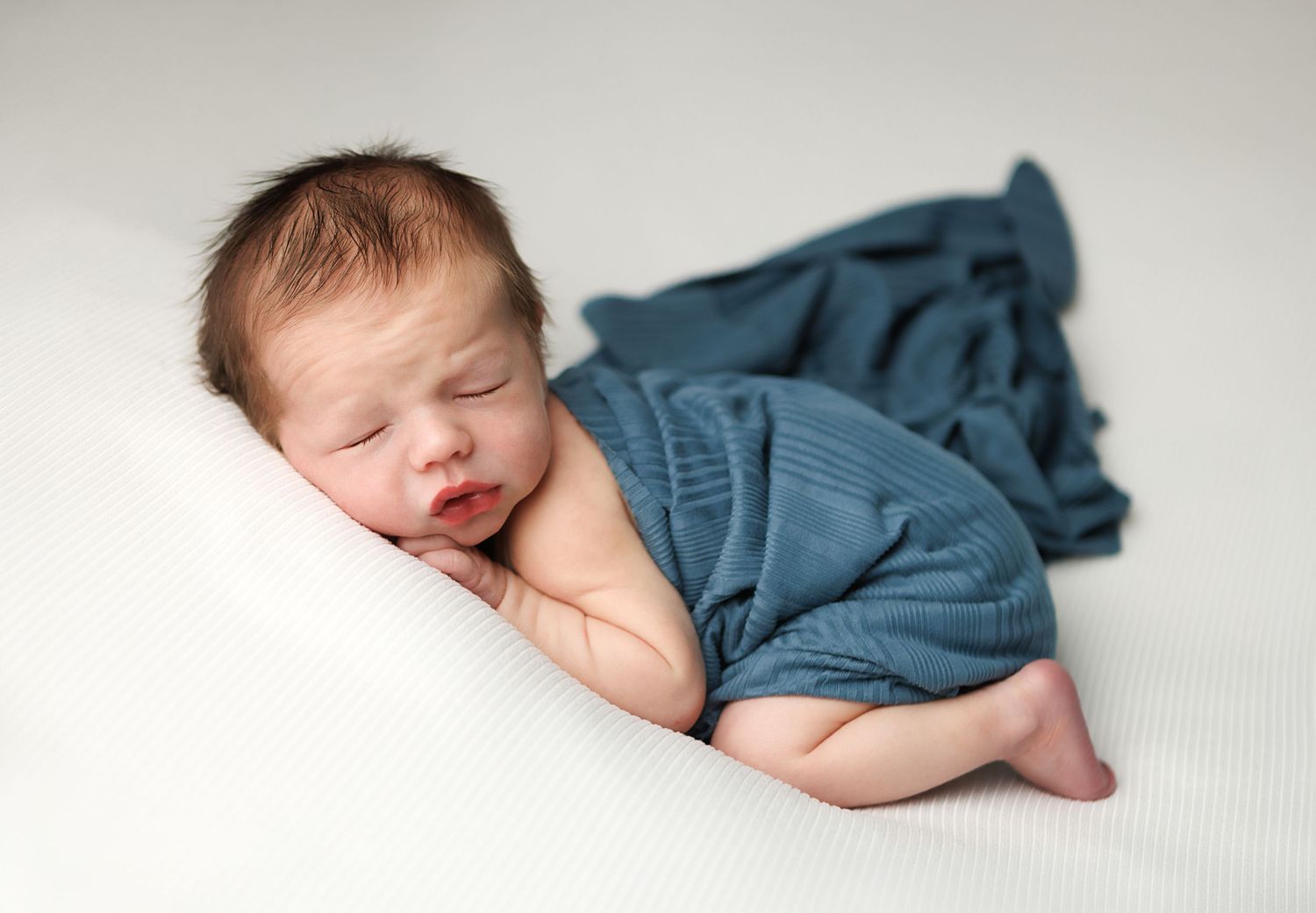 Peaceful newborn baby sleeping wrapped in a soft teal blue knit blanket on a white background.