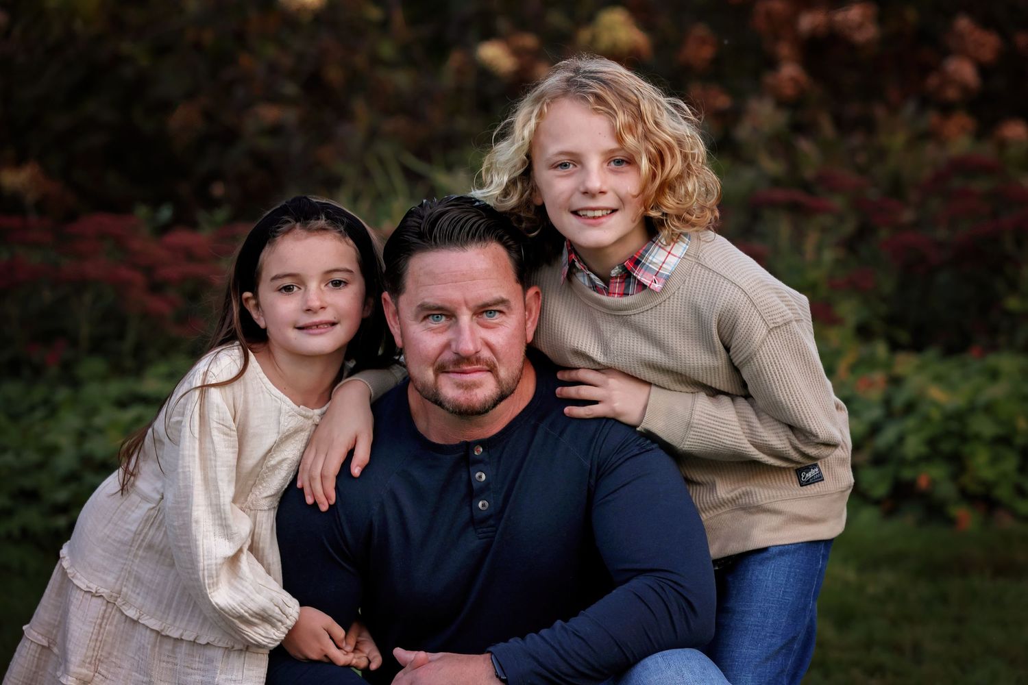 A family of three poses together outdoors in casual attire during an autumn photo session.
