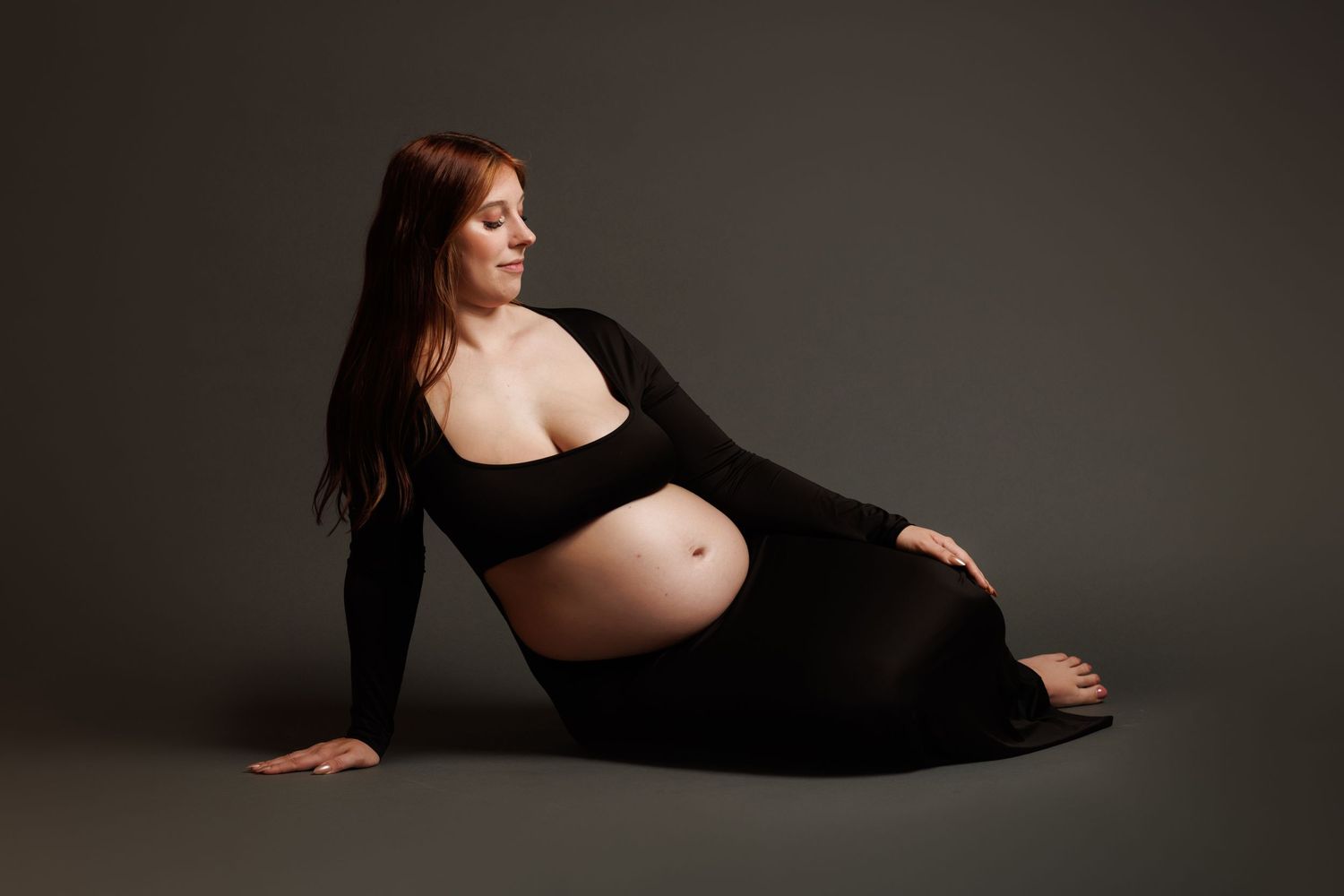 Pregnant woman in black dress posing gracefully on studio floor, showcasing baby bump in elegant maternity photography session.