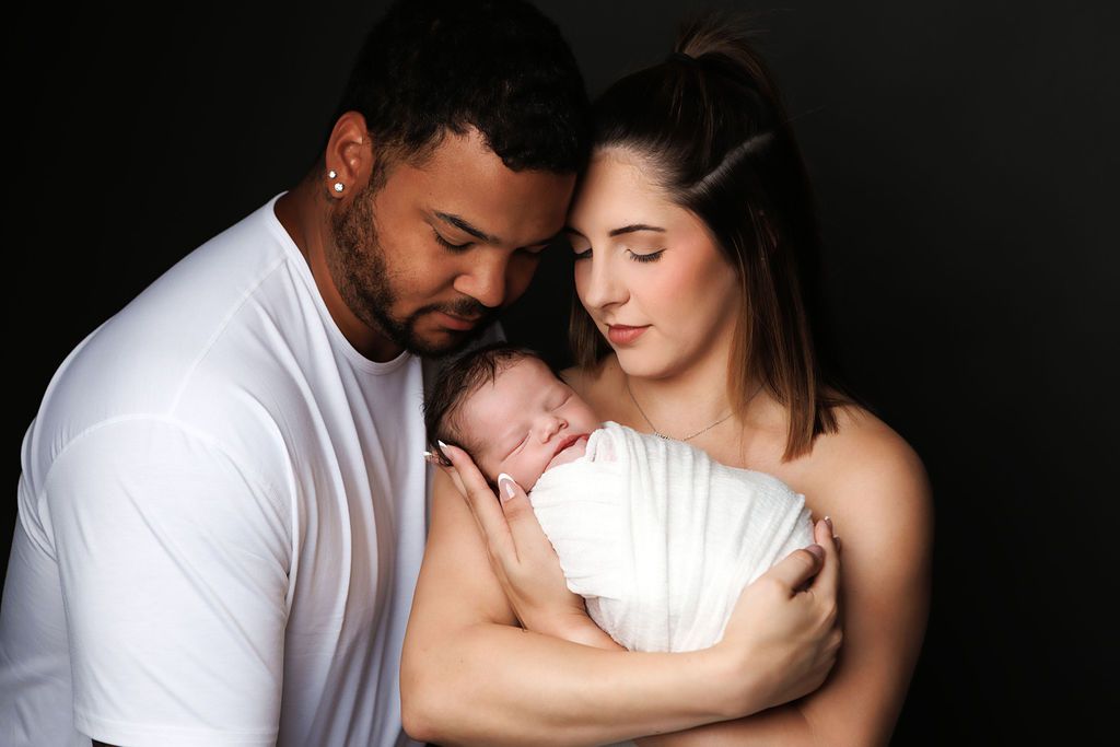 A couple tenderly holds their sleeping newborn baby wrapped in white against a dark studio background.