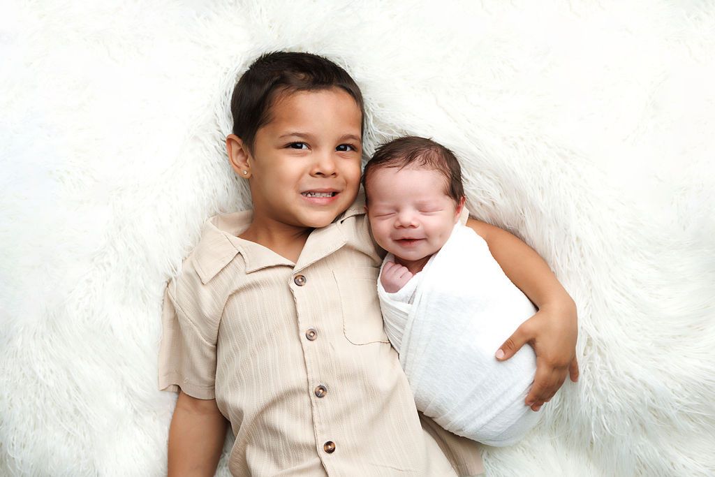 Young boy smiling while holding a swaddled newborn baby sibling on a white fluffy blanket.