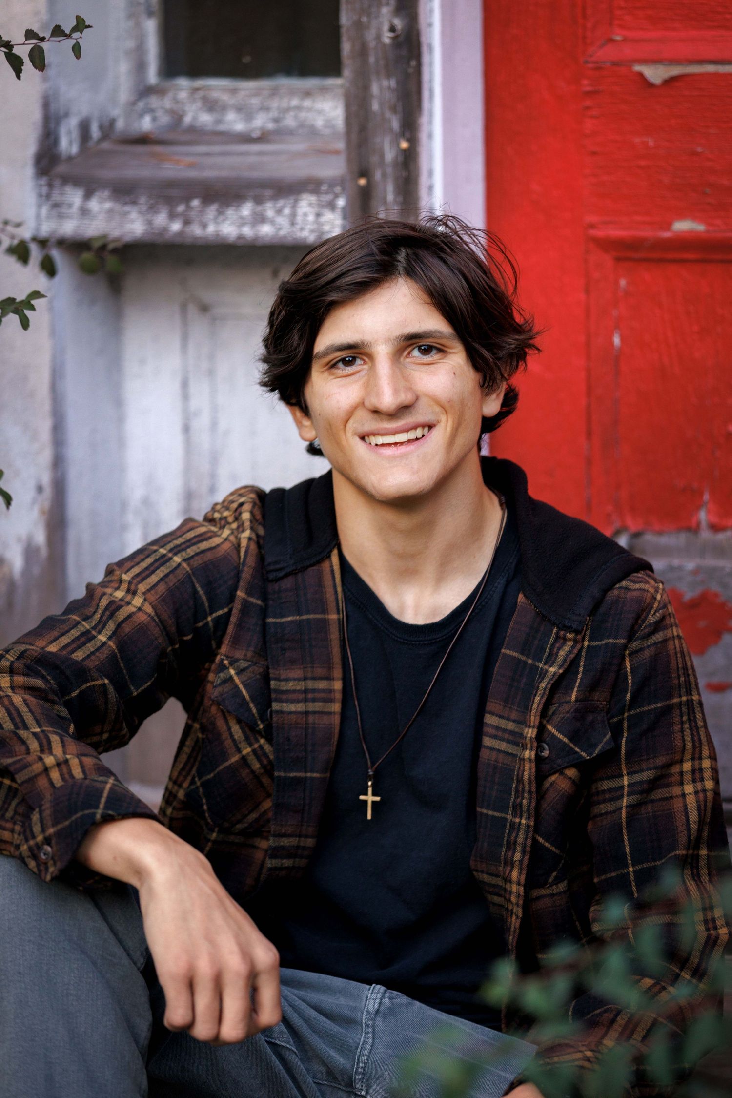 High School Senior in plaid flannel jacket and black t-shirt smiles while sitting against red door background.