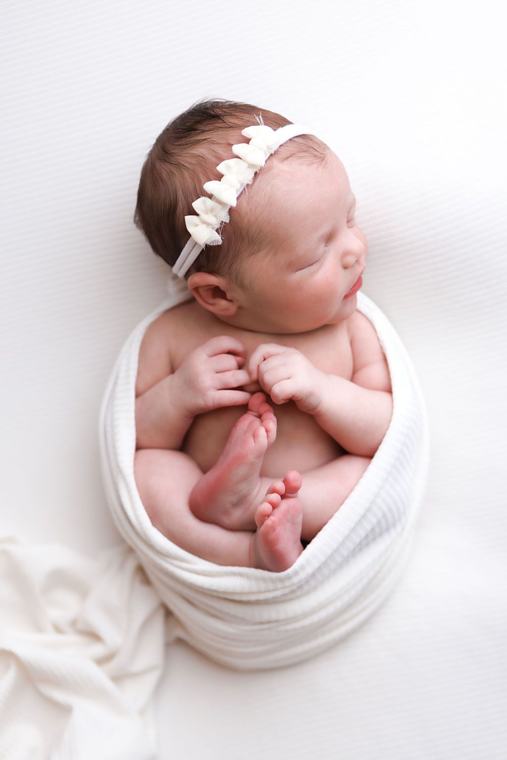 Sleeping newborn baby girl wrapped in white swaddle blanket with floral headband, peaceful studio portrait.