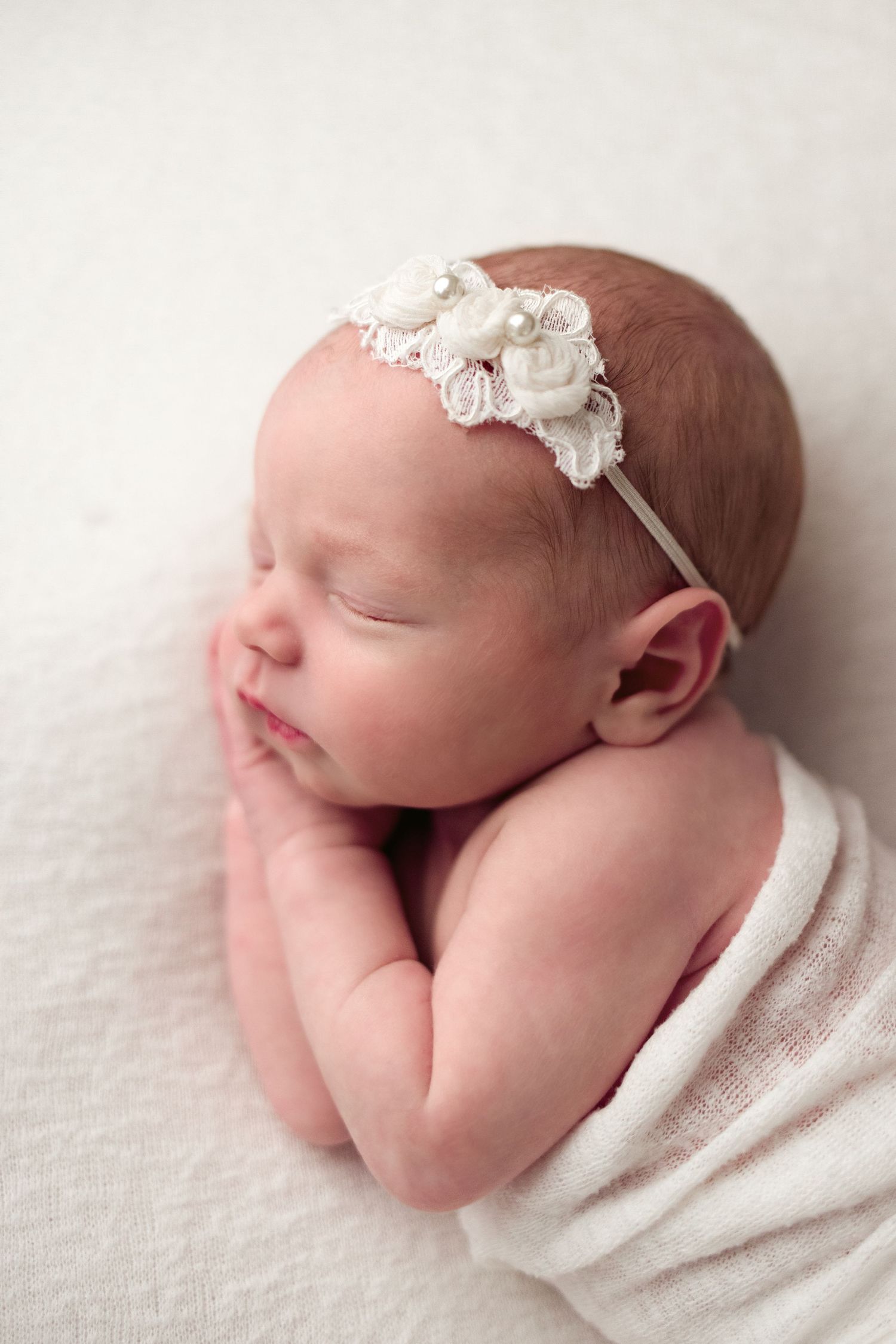 Sleeping newborn baby wrapped in white blanket wearing delicate lace headband during professional photo session.