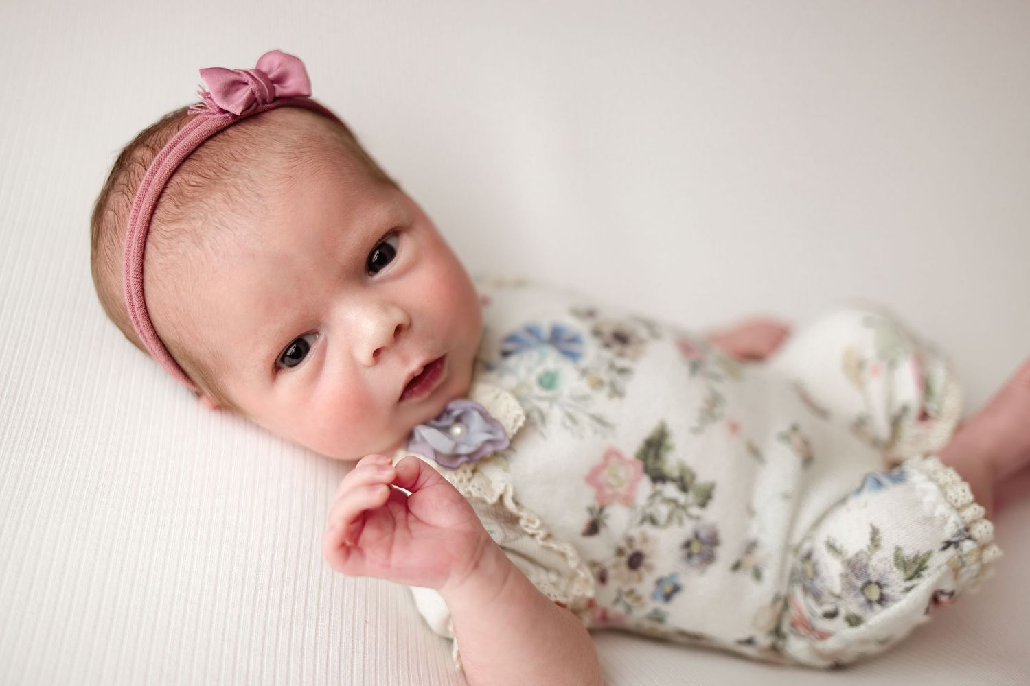 A baby wearing a pink bow headband and floral outfit lays on white bedding while looking at the camera.