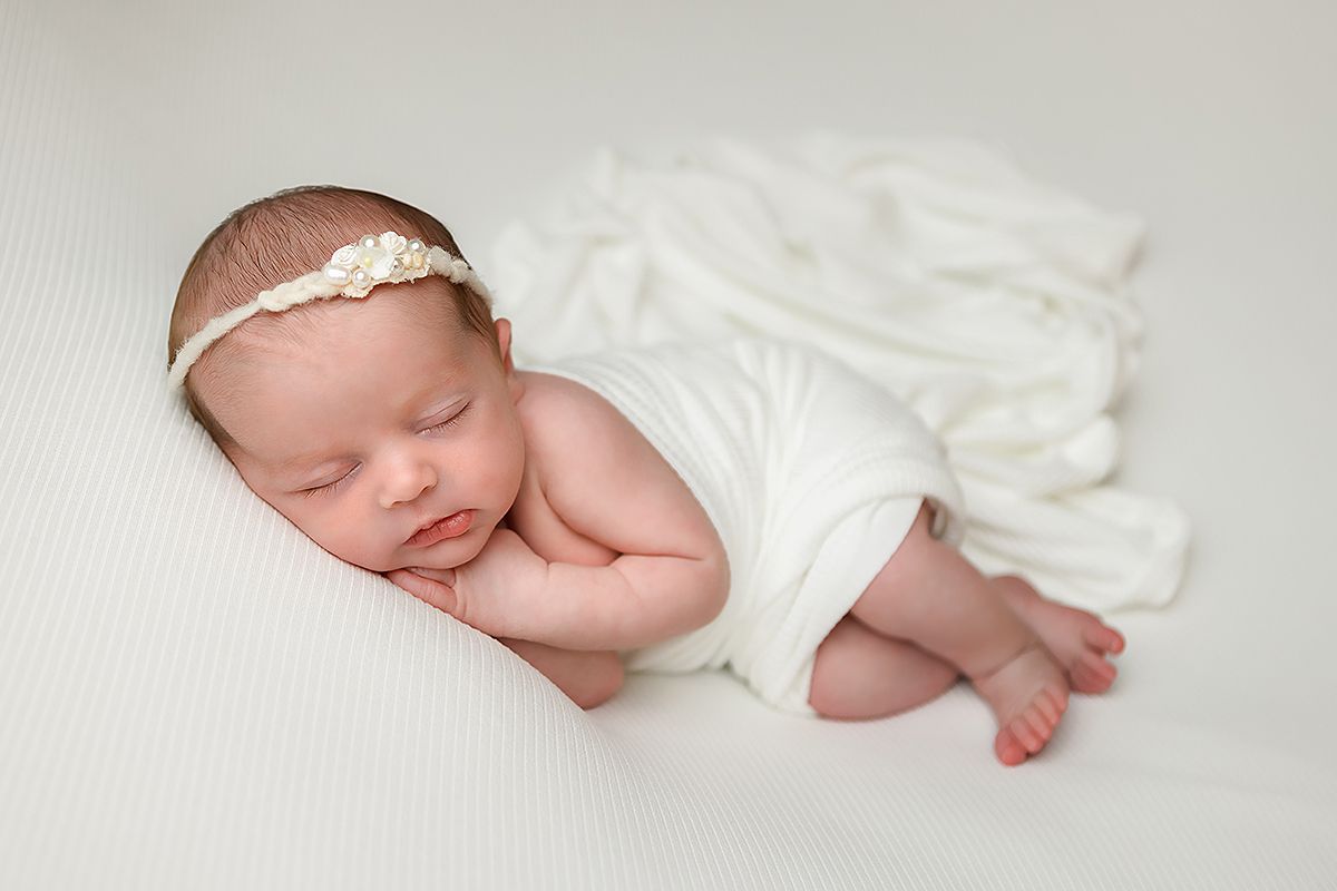 Newborn baby sleeping peacefully in soft white swaddle blanket and lace headband against neutral background.