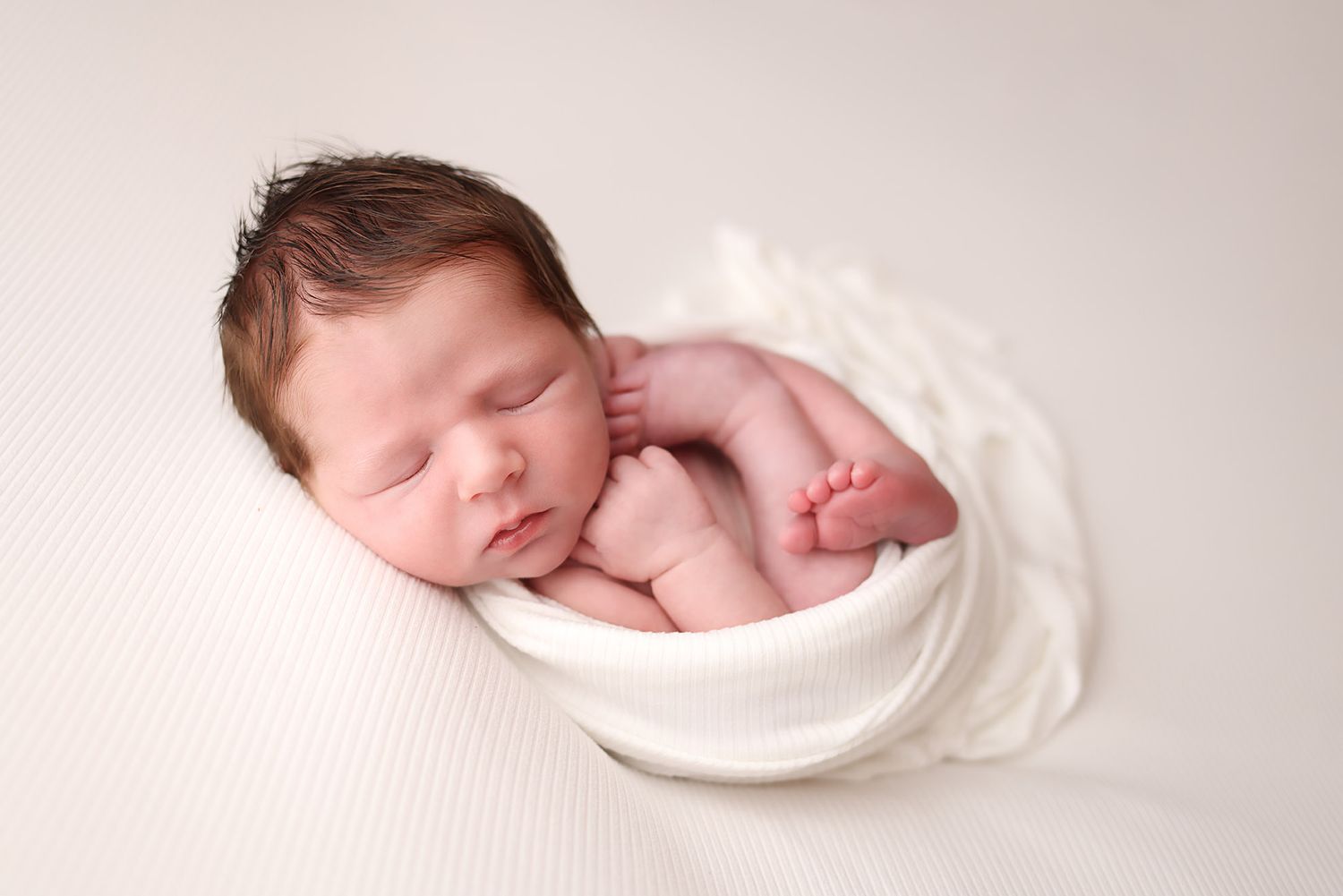 Newborn baby sleeping peacefully curled in a white bowl wrap on a soft cream background.