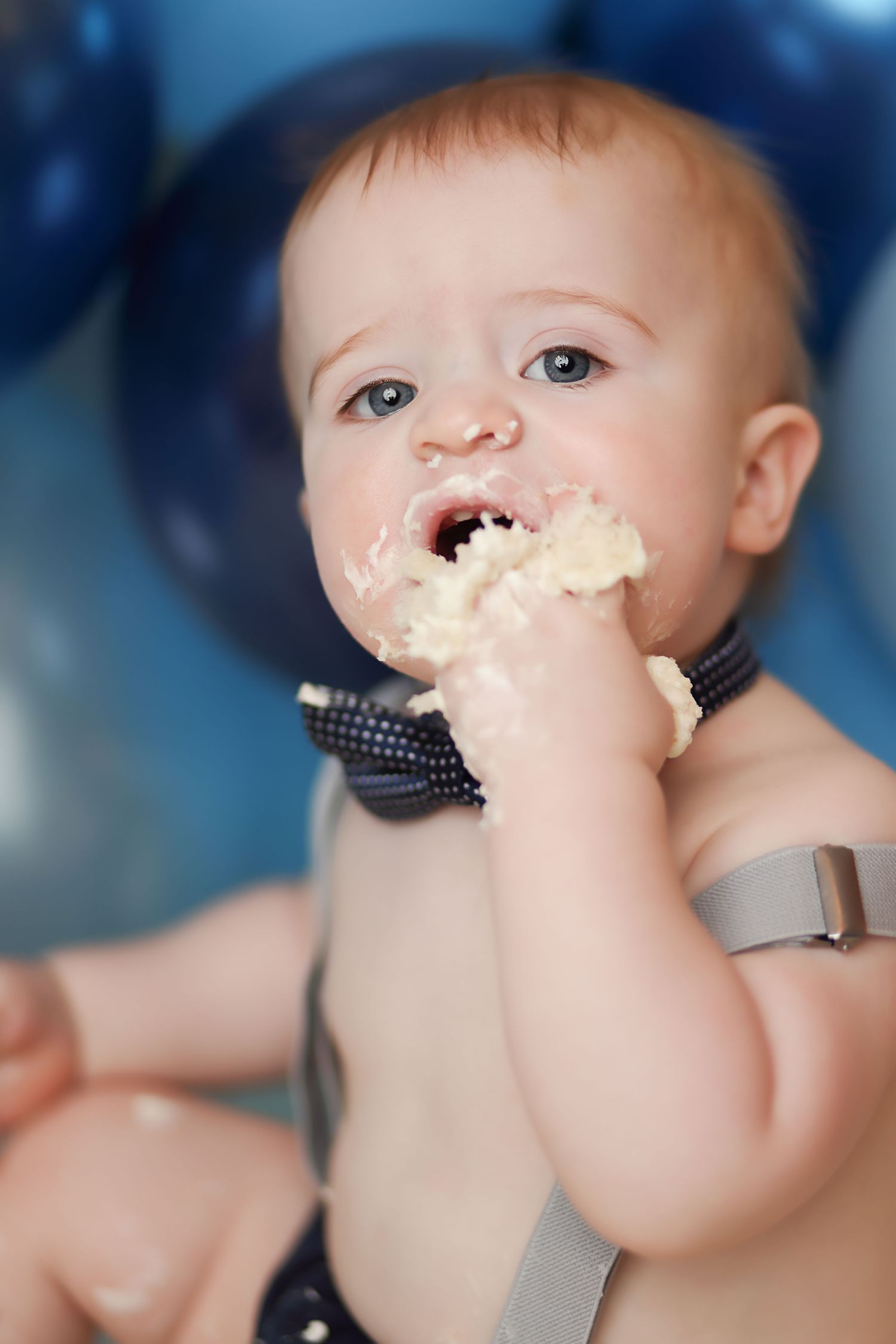 A baby with blue eyes eats birthday cake with hands during a first birthday smash cake celebration with blue balloons.
