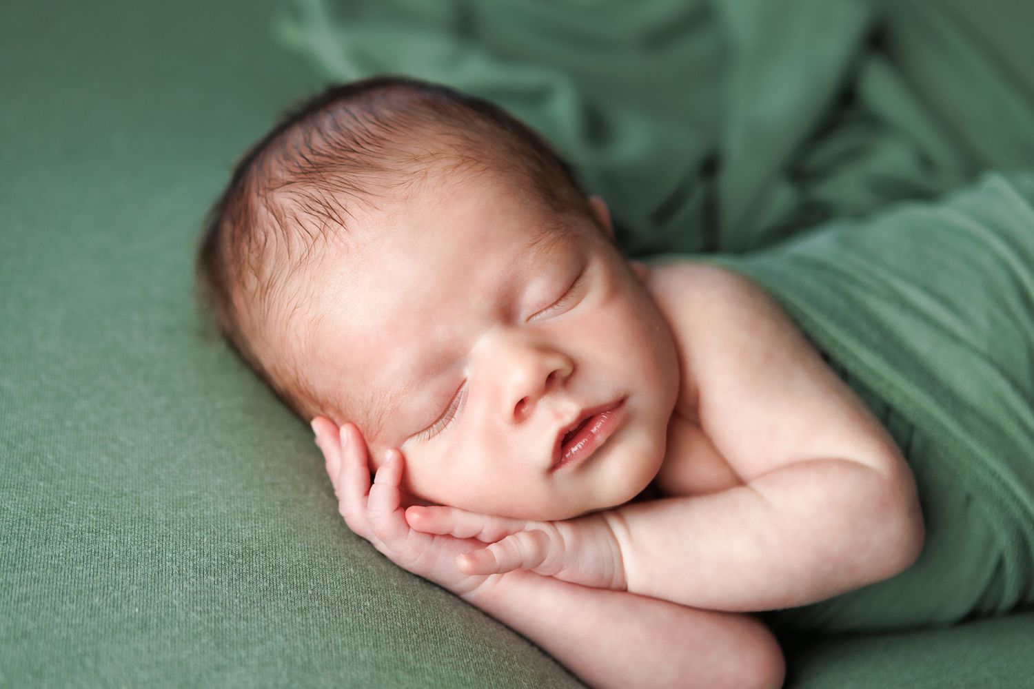 Peaceful newborn baby sleeping on green fabric, curled up with hands under cheek in professional photography.