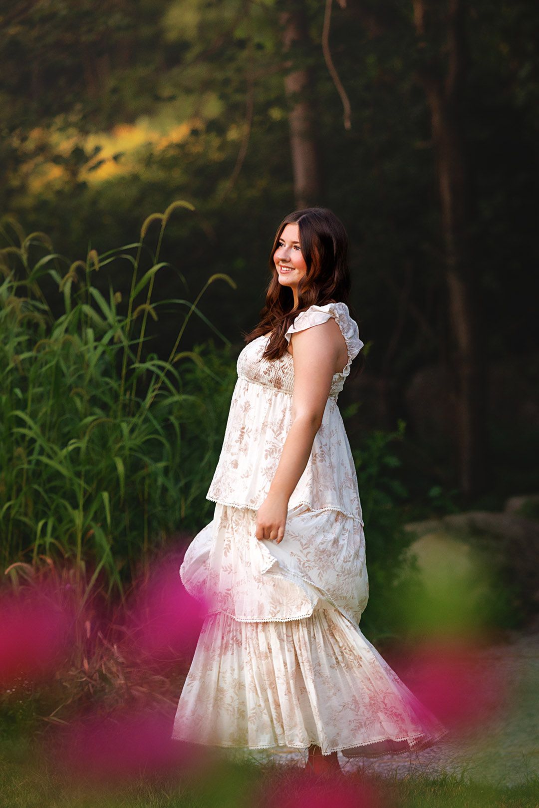 Young woman in elegant cream floral tiered gown smiling outdoors among lush greenery and pink flowers.
