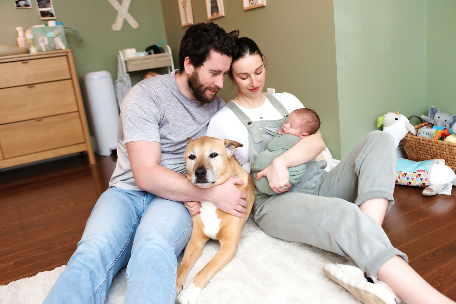 Young family with newborn baby and golden dog relaxing together on white rug in cozy nursery room.