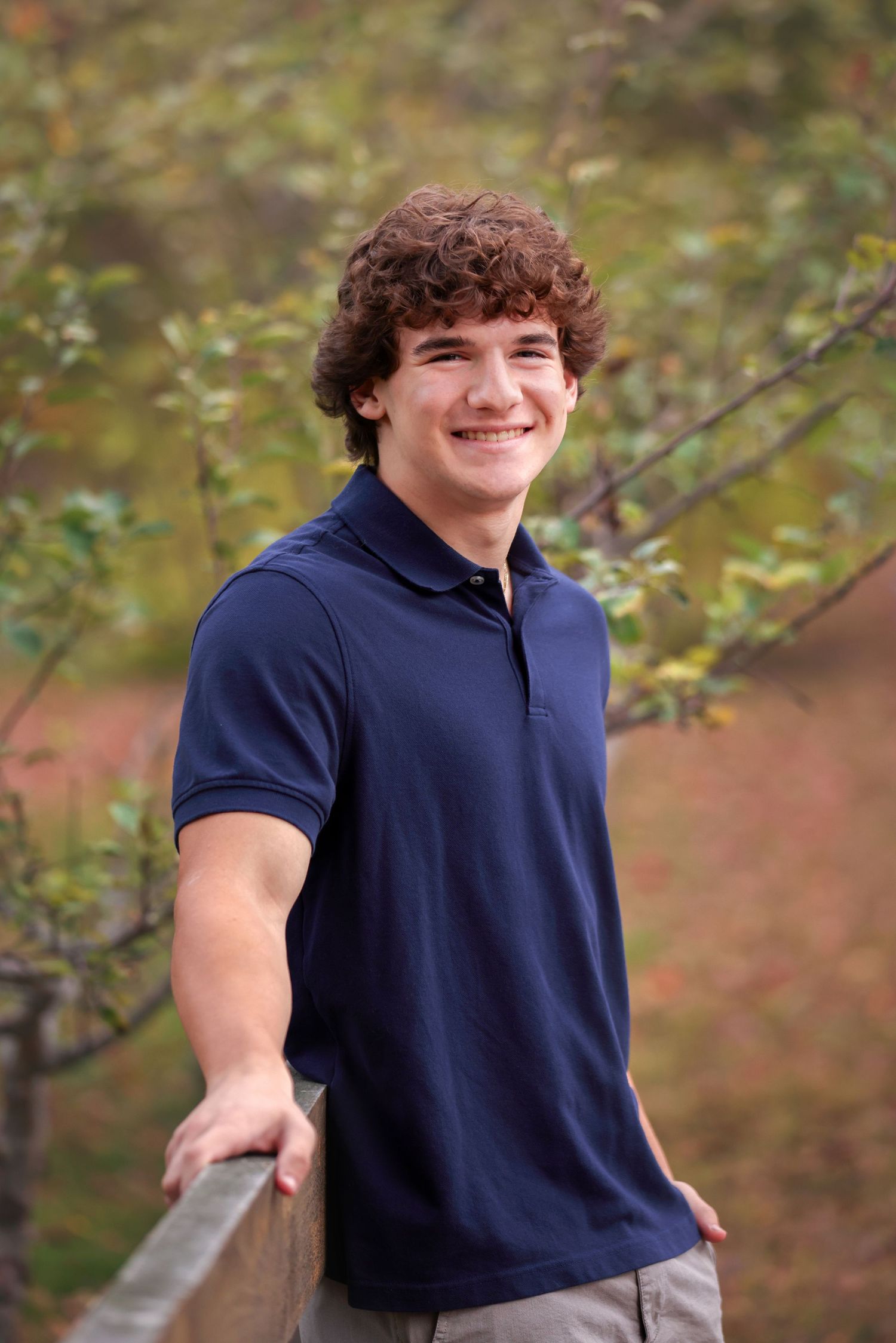 High School Senior in a navy blue polo shirt smiles while standing outdoors against a blurred natural background.