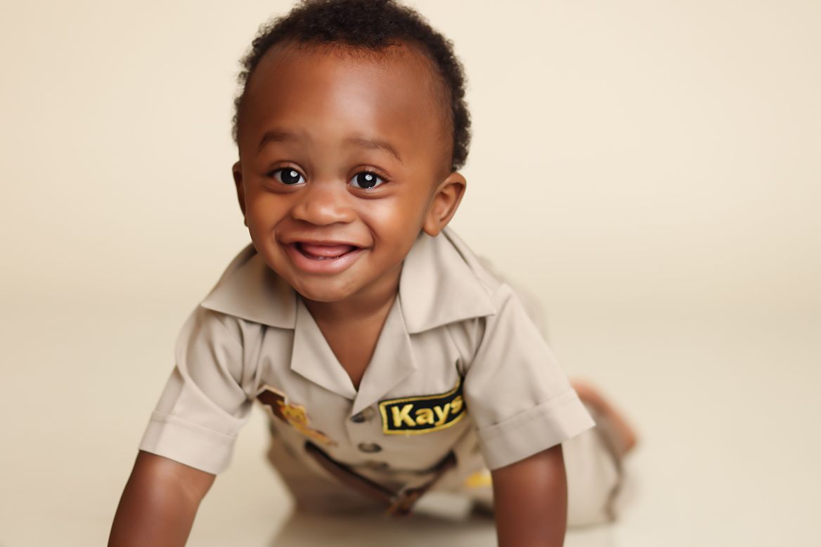 A young child wearing a beige collared outfit smiles brightly while sitting against a light-colored background.