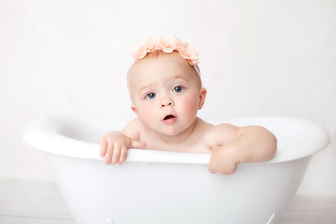 Sweet baby peeking over edge of white bathtub during bath time with innocent expression. Boston Cake Smash Session.