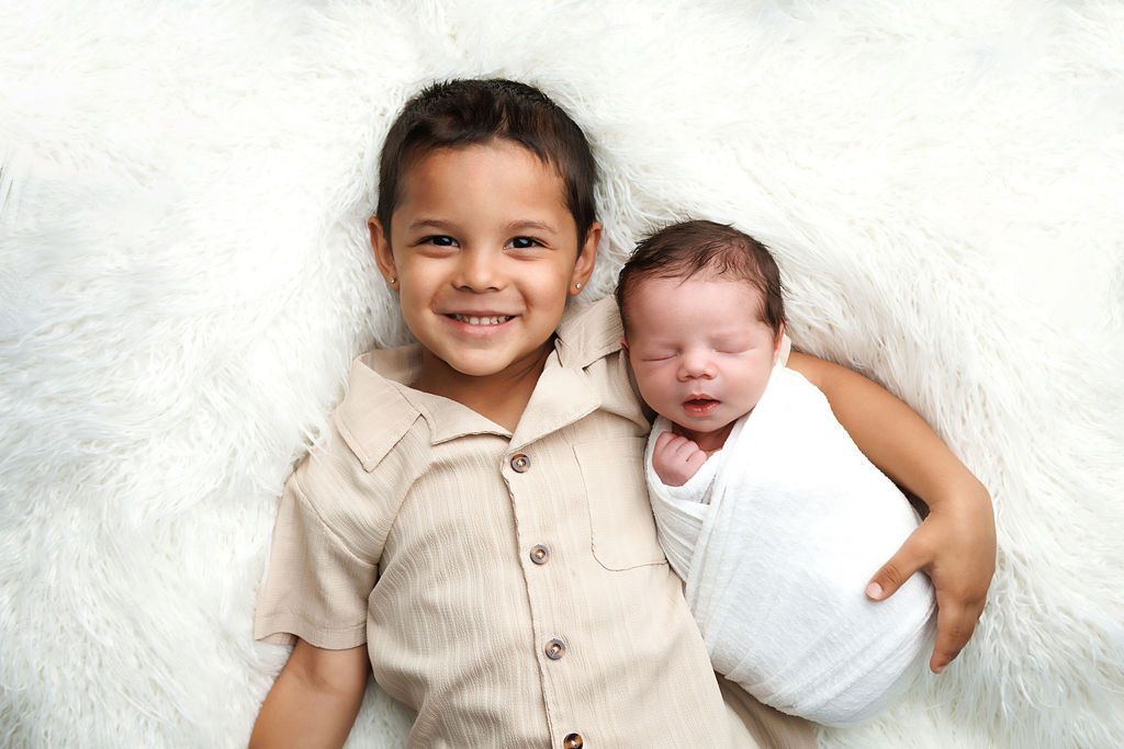 Smiling young boy lying on white fluffy rug with sleeping newborn baby sibling wrapped in white swaddle.