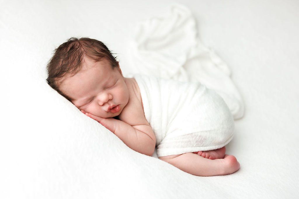 Peaceful newborn baby wrapped in white swaddle sleeping on soft white blanket in professional photo.