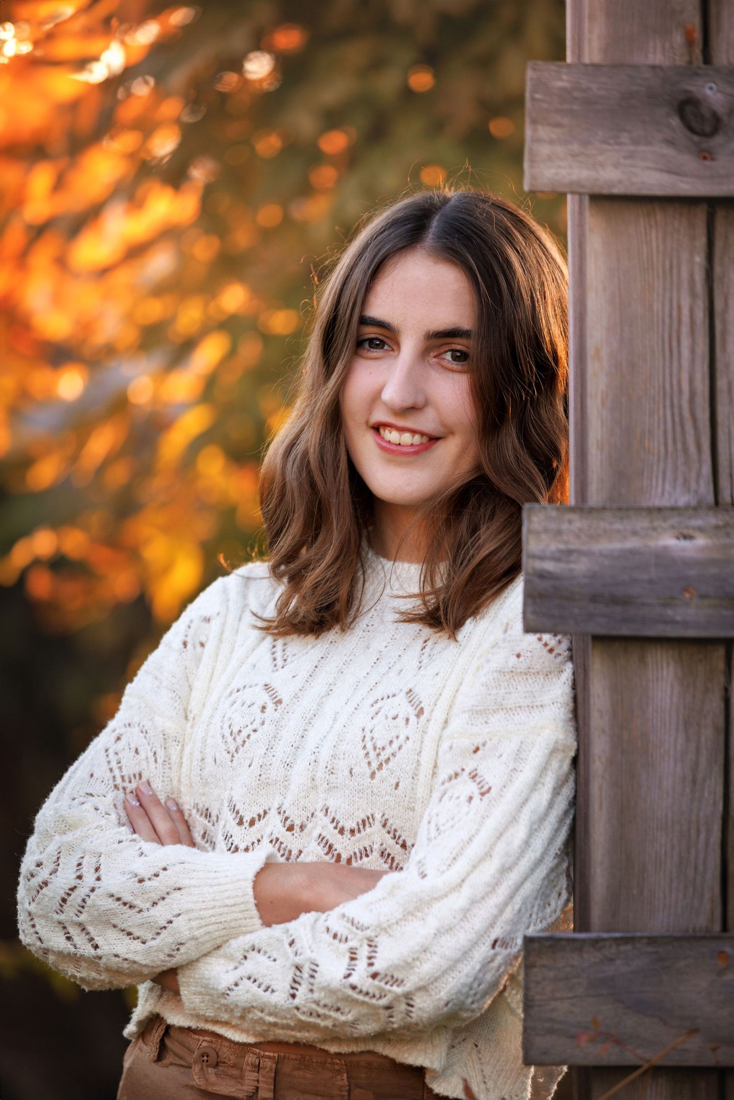 A high school Senior in a white knit sweater poses with crossed arms against a wooden post with autumn colors in the background.