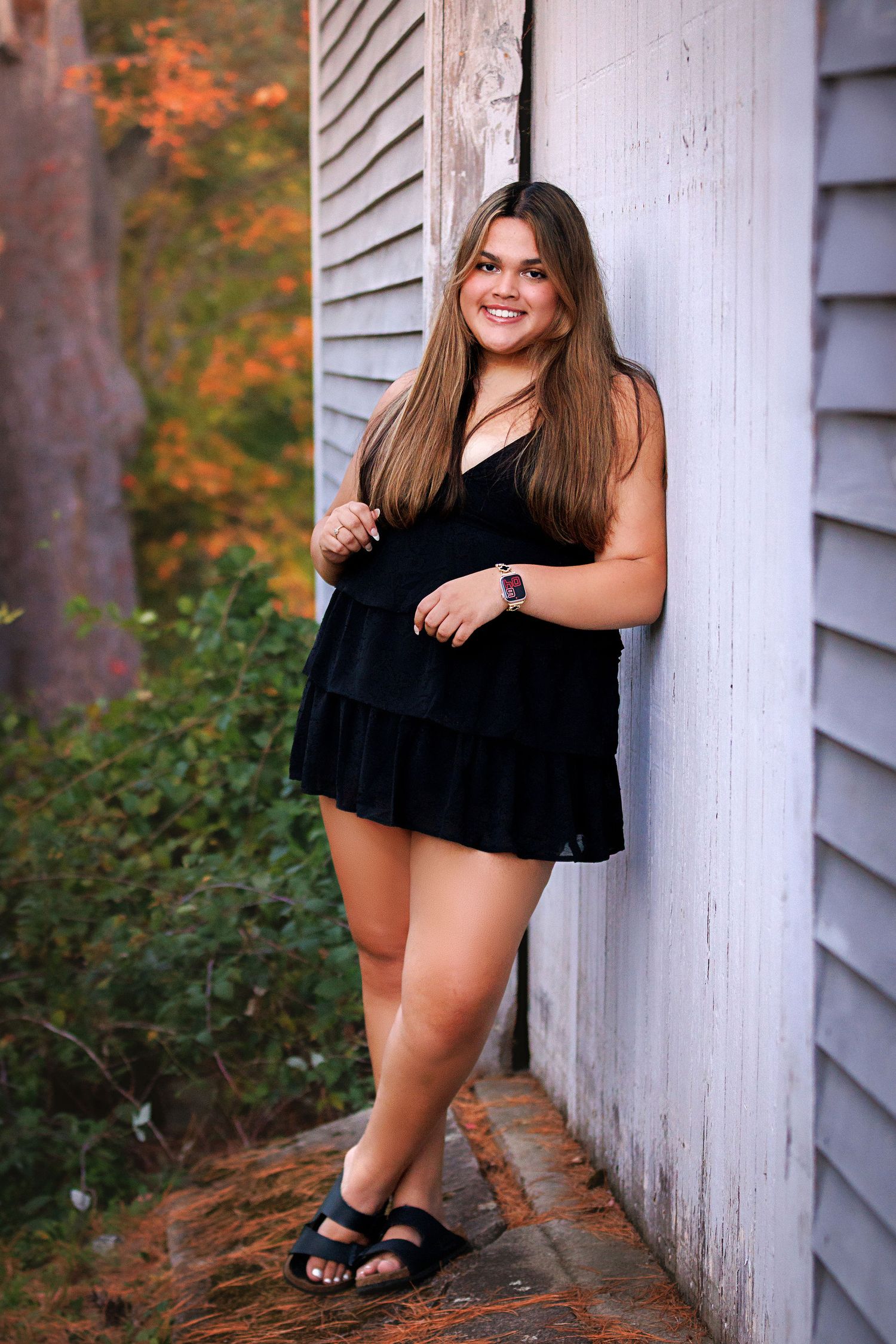 Young woman in black mini dress and sandals smiling against white wall with autumn foliage background.