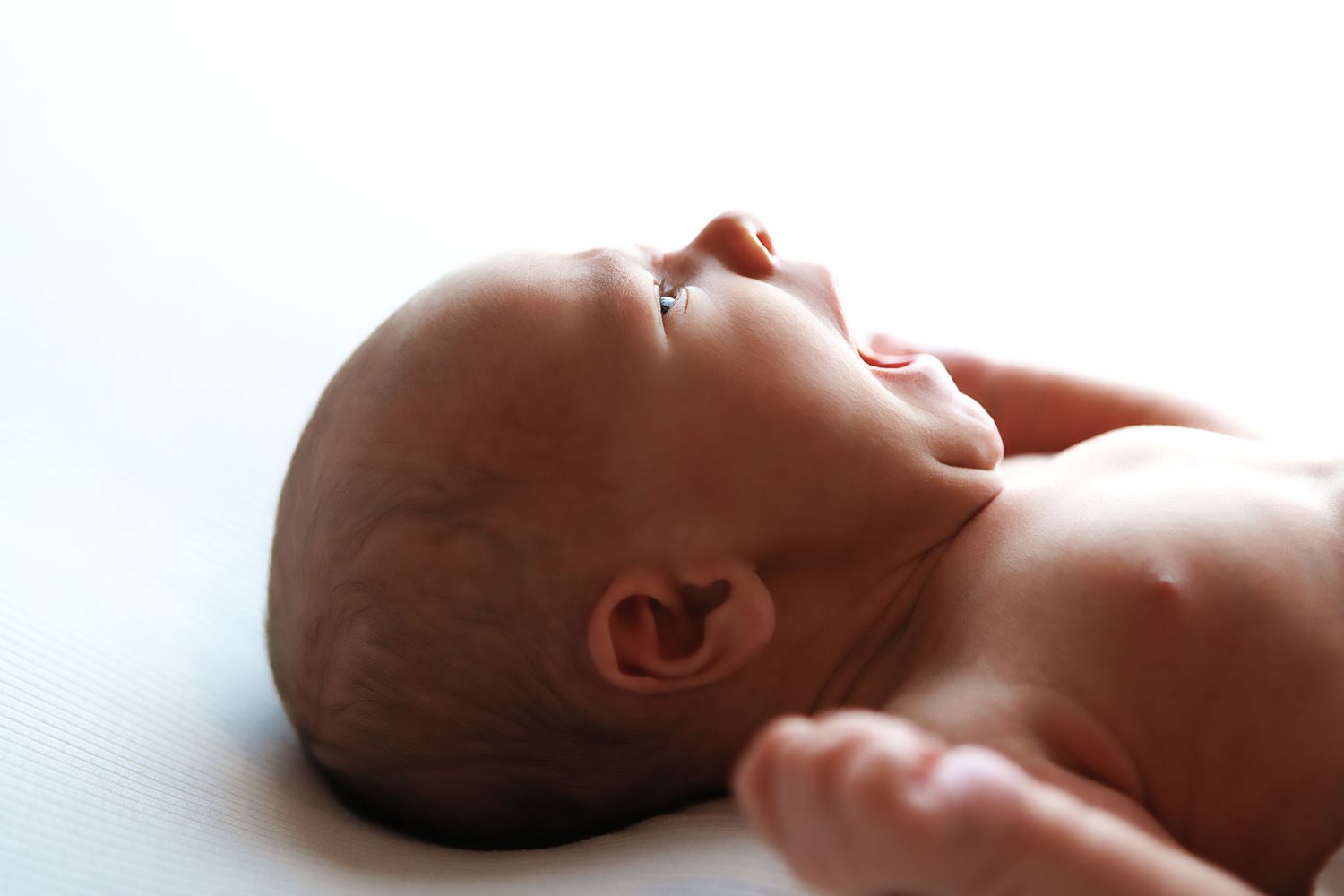 Peaceful newborn baby lying on back with eyes closed, profile view showing delicate features on white background.