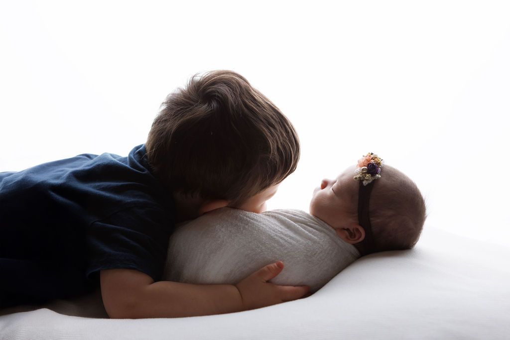 Young boy gently kisses newborn baby sibling wrapped in white swaddle on white background.