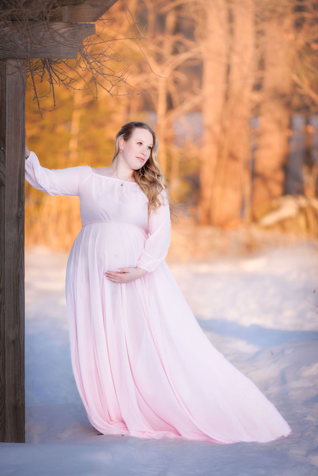 Pregnant woman in flowing pink gown poses gracefully in snowy winter landscape at golden hour.