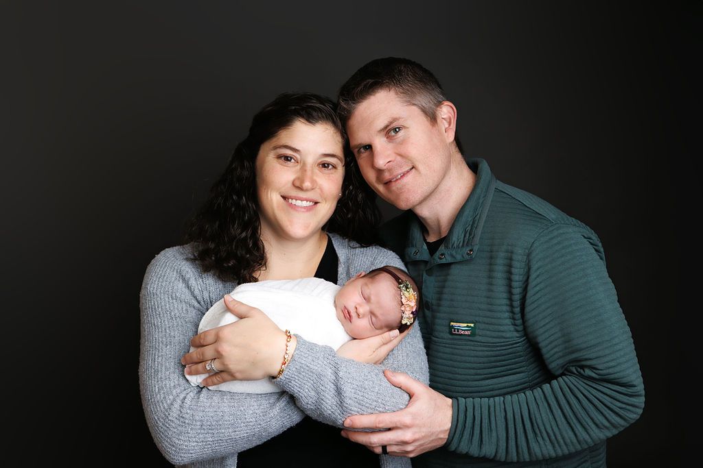 A smiling couple poses with their newborn baby wrapped in white against a dark studio background.