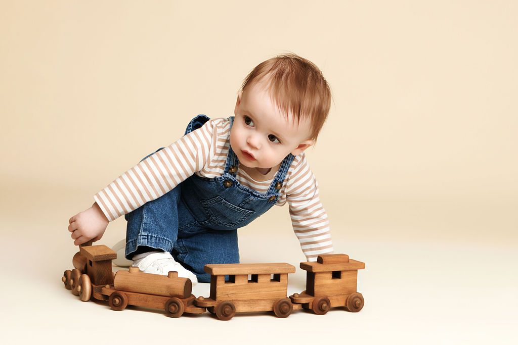 Baby in denim overalls playing with a wooden toy train set on a beige background.