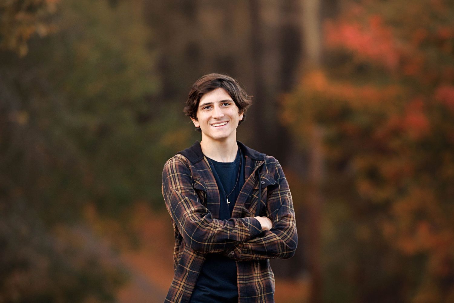 Young man with crossed arms smiling outdoors in autumn forest wearing plaid shirt, fall foliage background.