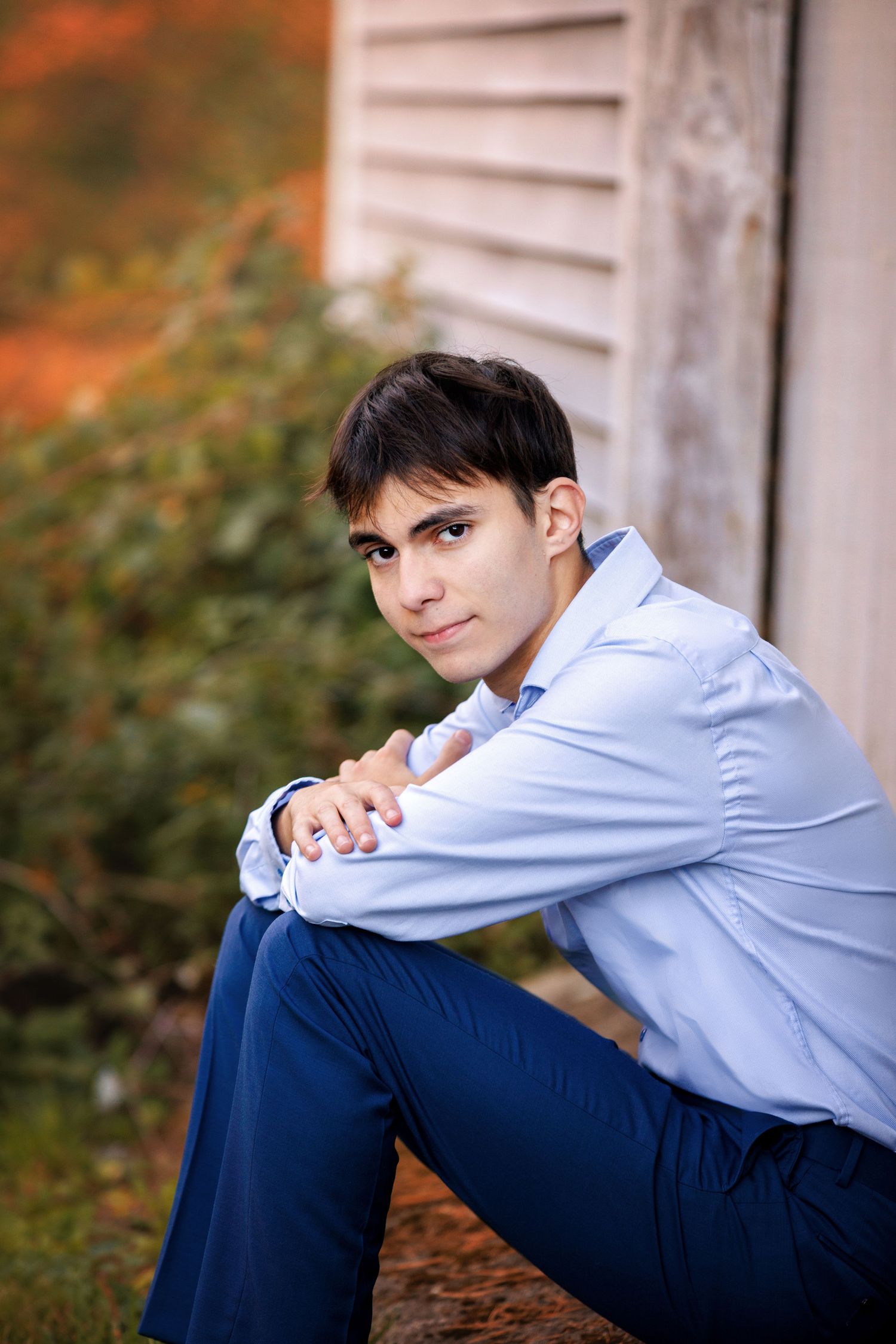 Young man in light blue shirt and navy pants sits outdoors, arms resting on knees, with greenery and building behind him.