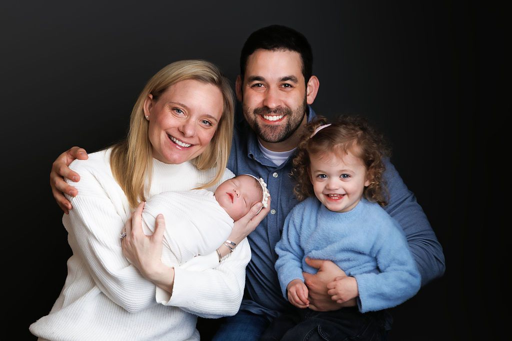 Happy family portrait with parents holding newborn baby and smiling toddler girl against dark background.