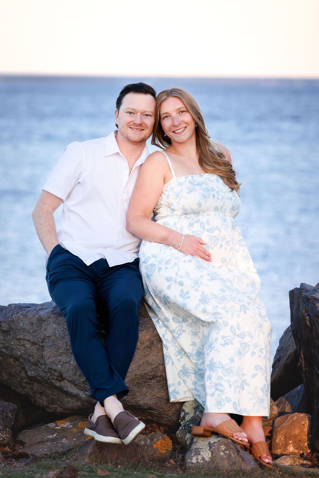 Happy pregnant couple sitting on coastal rocks at dusk, woman in blue floral sundress.