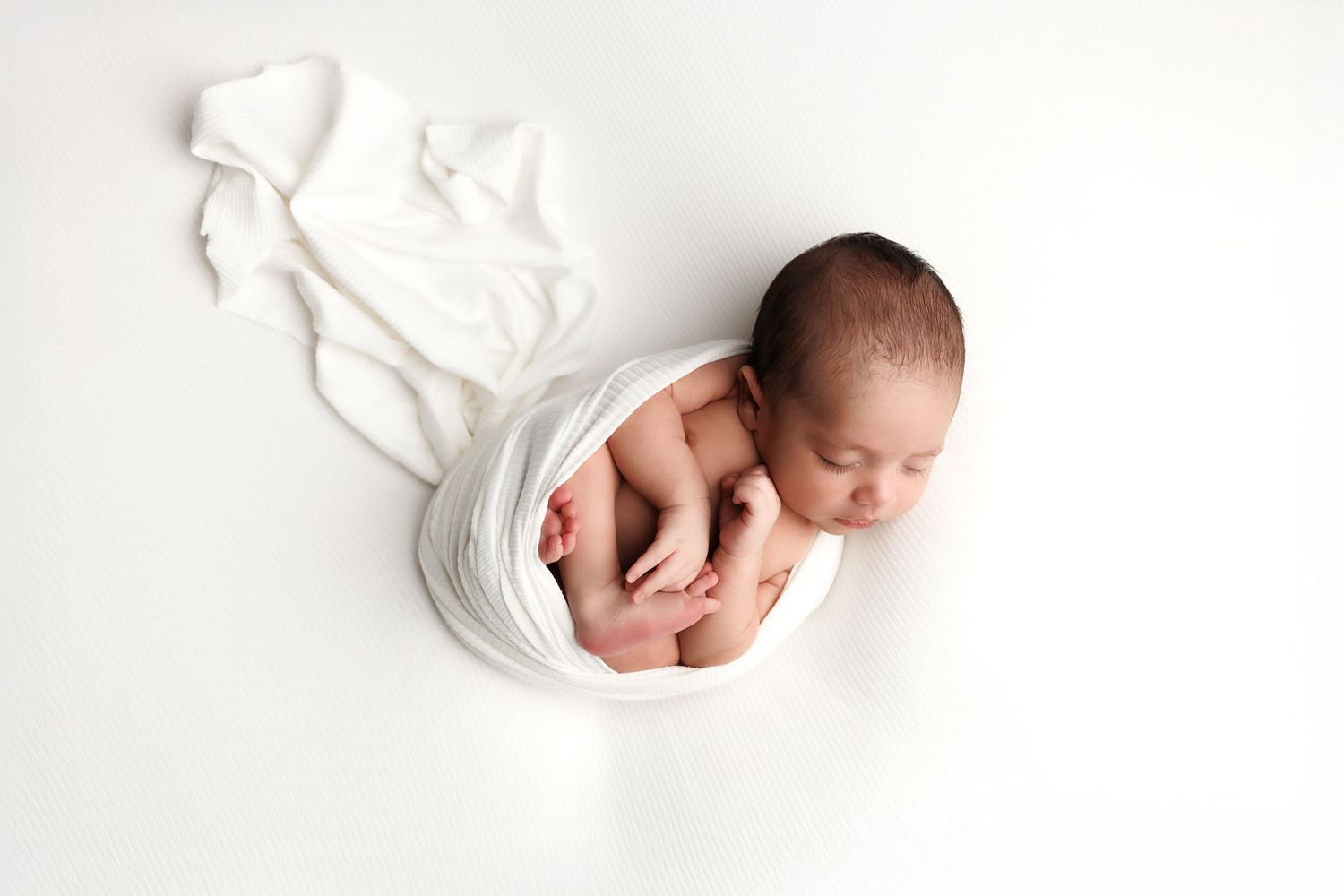 A sleeping newborn baby lies on white fabric with delicate hands visible in the frame.