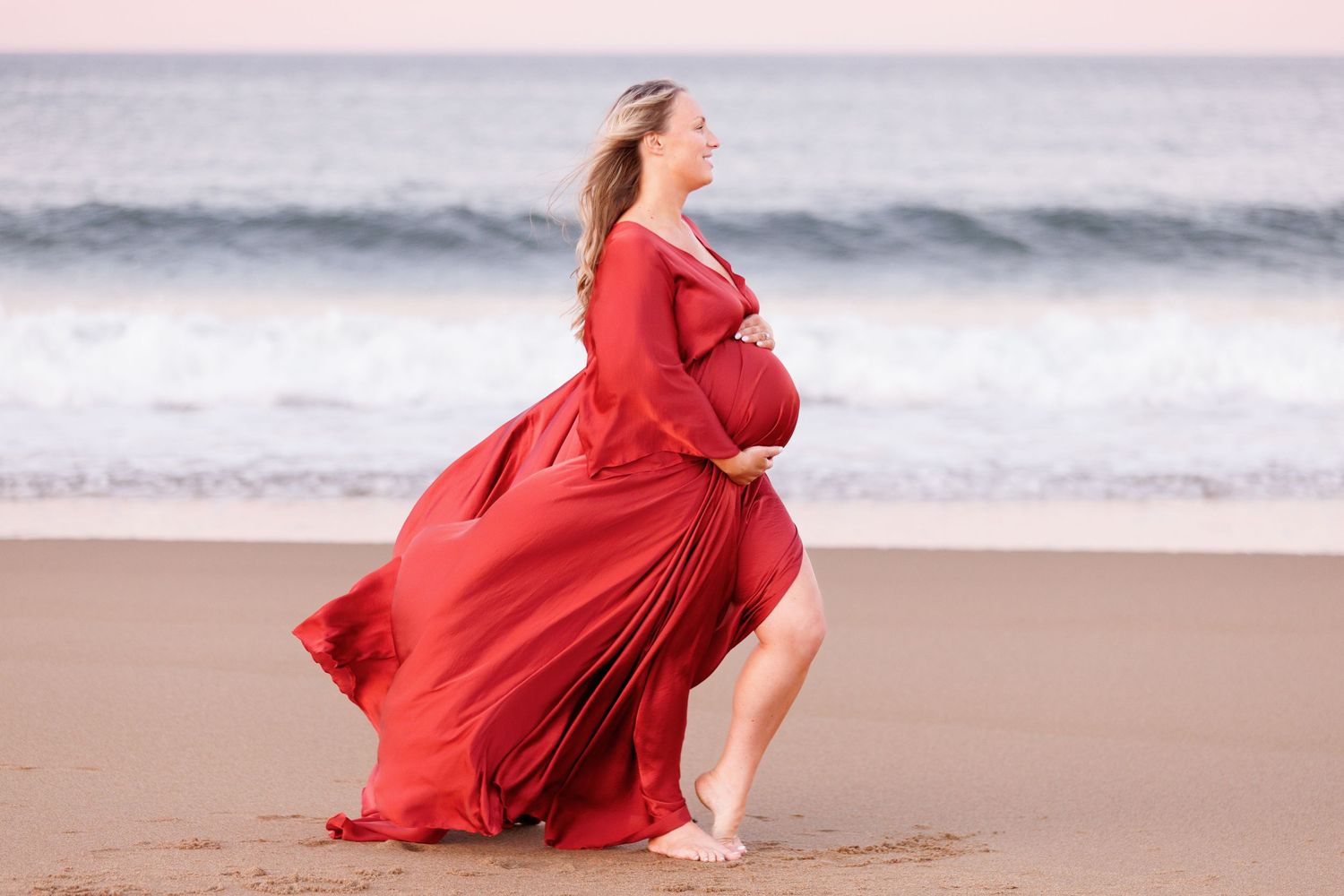 Pregnant woman in flowing red gown walking barefoot on sandy beach with ocean waves in background.