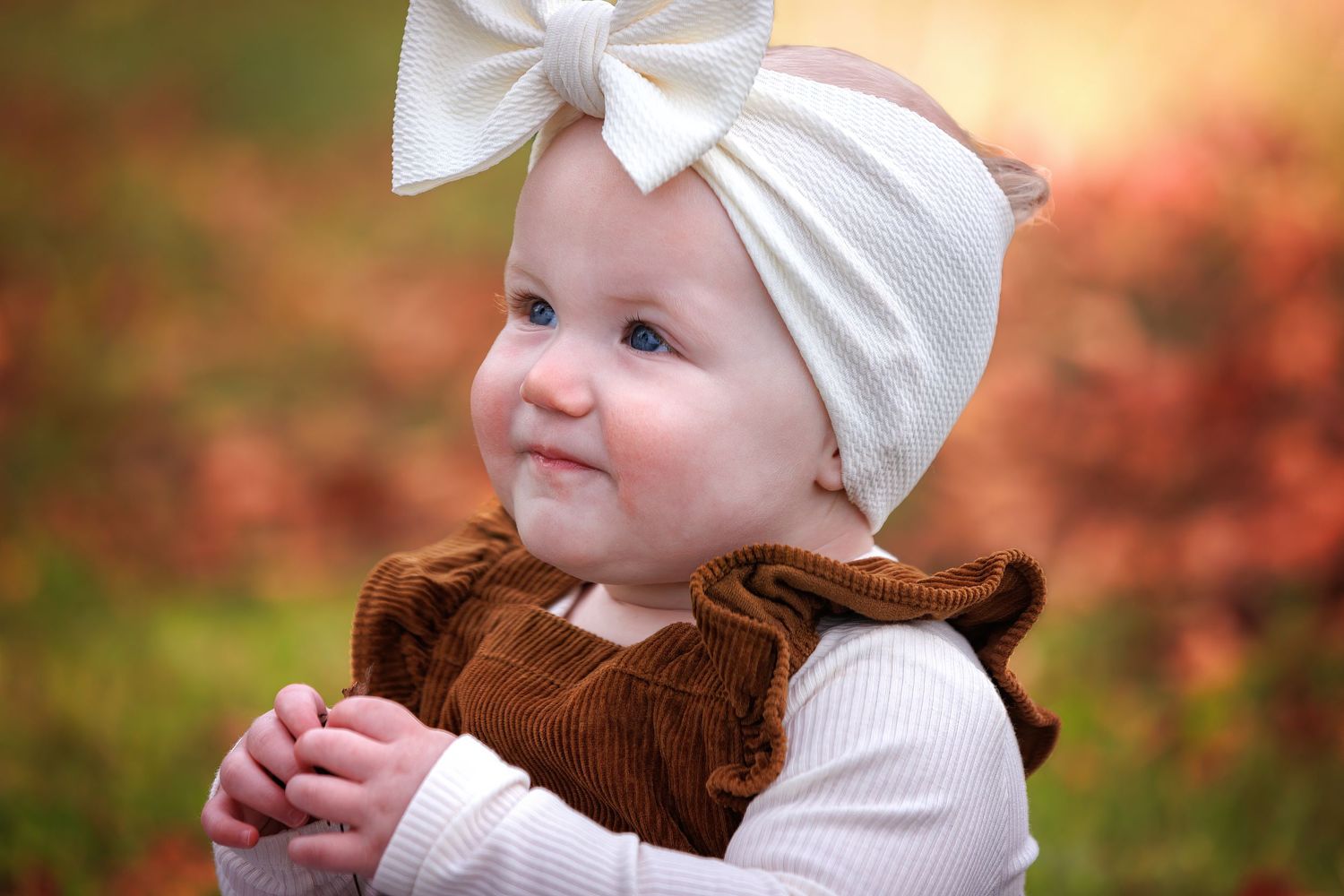 A baby wearing a white bow headband smiles while sitting outdoors with autumn foliage in the background.