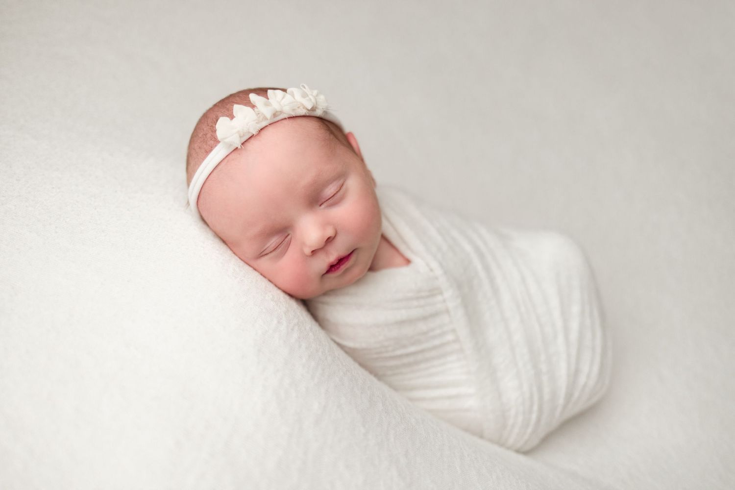 A sleeping newborn baby wrapped in white swaddle blanket lies peacefully on white bedding.