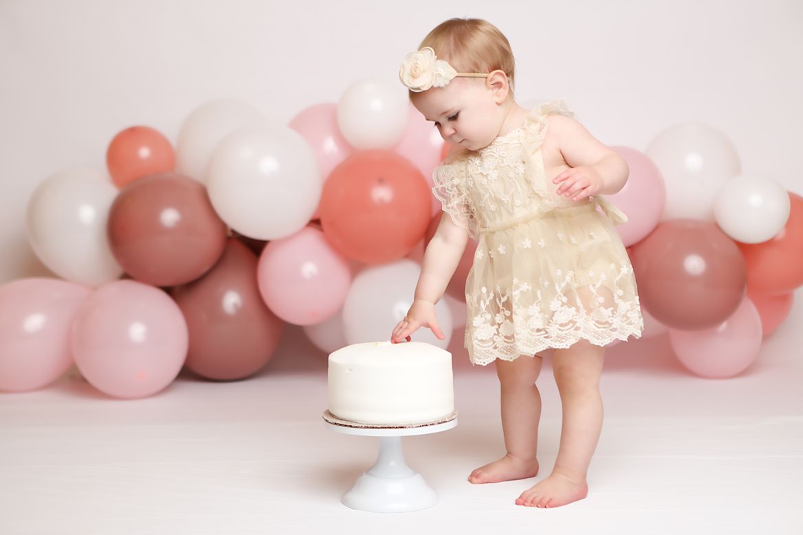 Baby in pink dress exploring white studio setup with pink and white balloon decorations.