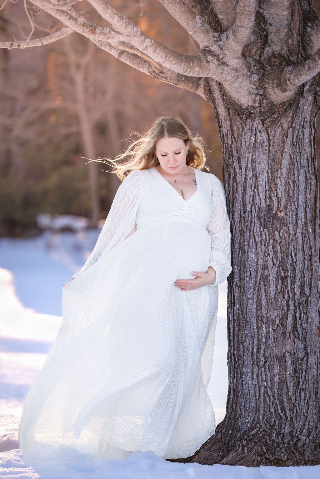 Pregnant woman in white lace gown leaning against a tree in a snowy winter landscape.