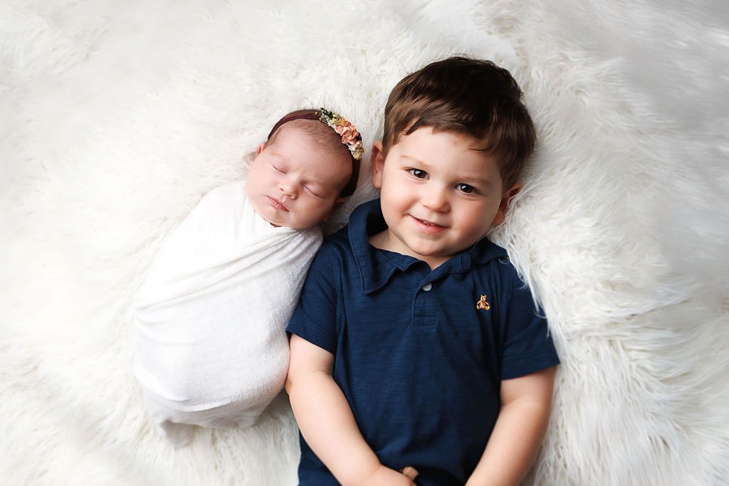Smiling toddler boy in navy polo lying on white fur with sleeping swaddled newborn baby sister wearing floral headband.