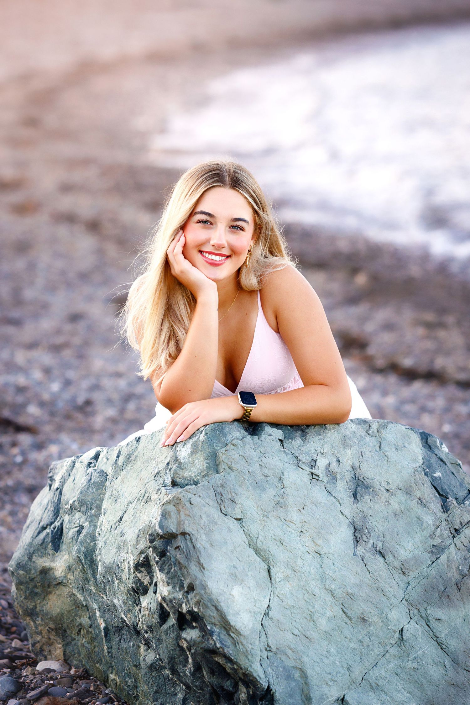 A person in white poses on a large blue-gray rock formation at the beach.