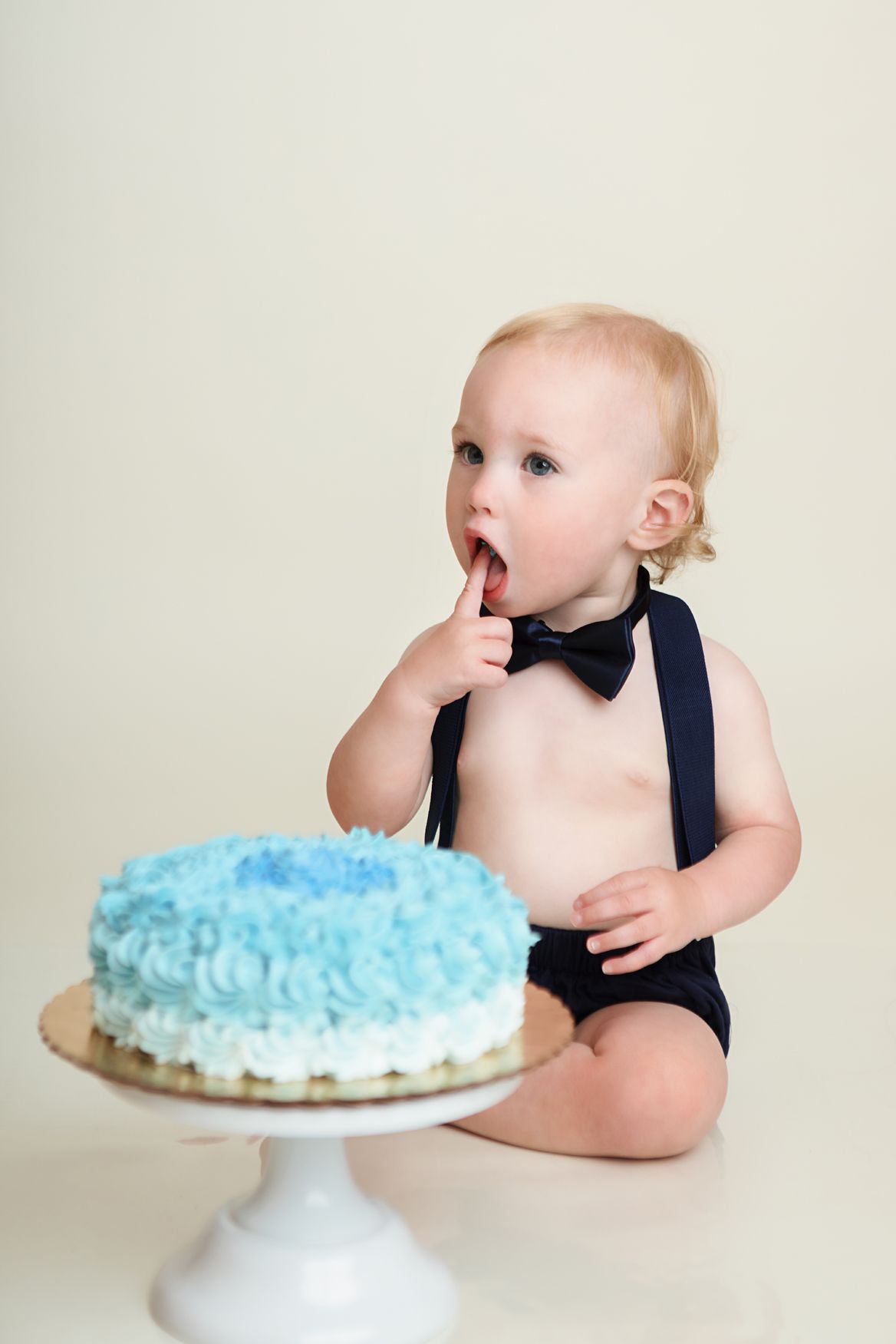 A sequence of photos showing a baby in navy blue overalls reaching for and reacting to a blue-frosted cake on a white stand.