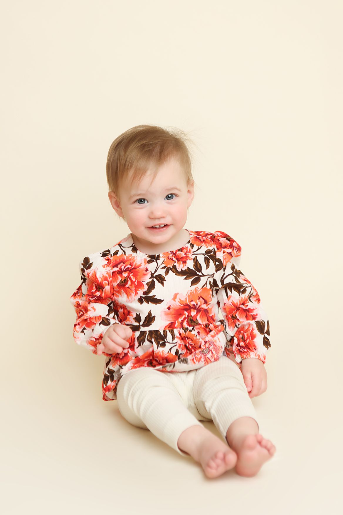 Baby in floral outfit with coral and white pattern lies on cream background showing bare feet.